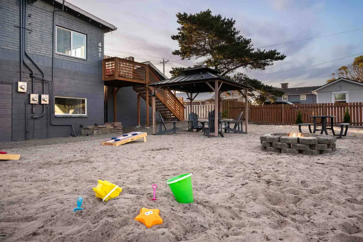 A cheerful exterior scene featuring a gazebo, fire pit, gravel pathway, and playful beach toys in the sand. Children can be seen playing while adults gather under the gazebo, creating an inviting space for both relaxation and social interaction.