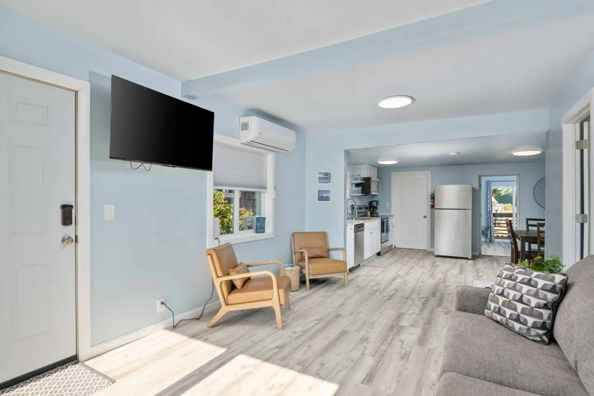 A cozy living area with a comfortable seating arrangement is bathed in natural light from a large window. Modern kitchen appliances can be seen in the background, seamlessly blending the living and kitchen spaces. The mood is inviting, with soft furnishings and a sense of openness, perfect for relaxing or socializing.