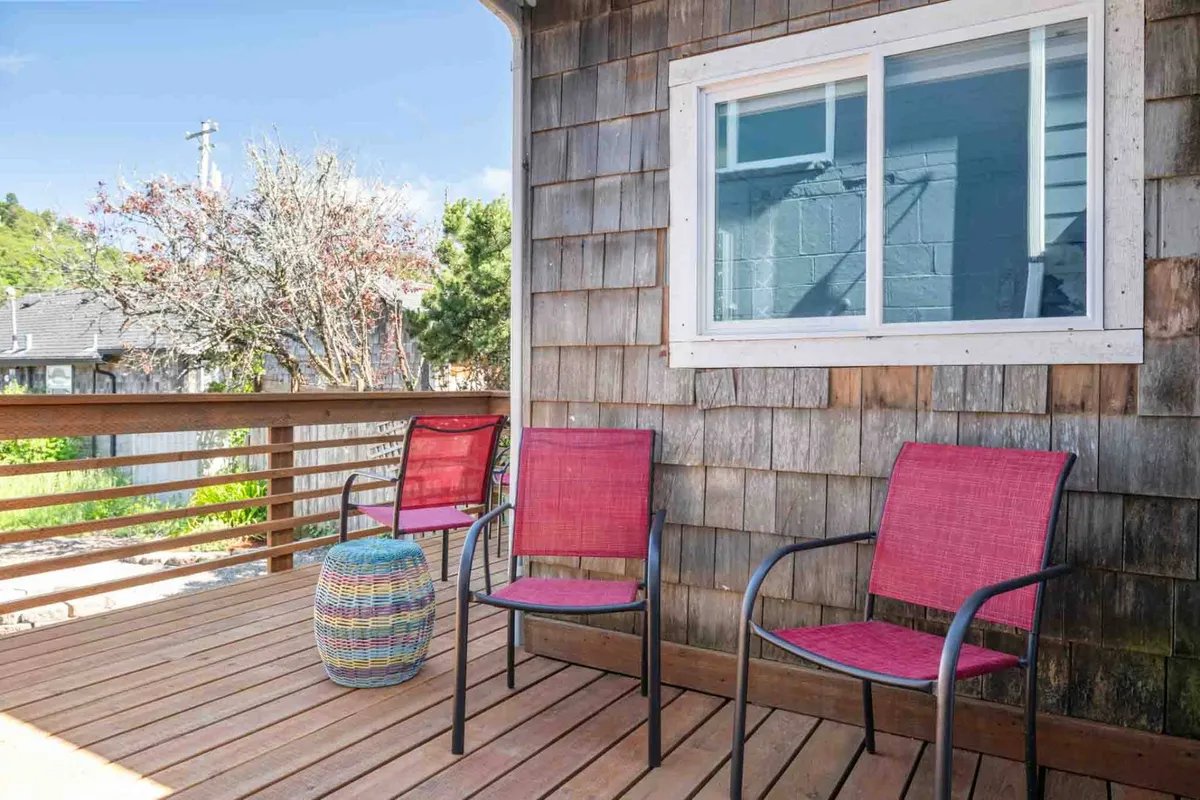 A sunny outdoor deck with two red chairs and a colorful decorative table provides a comfortable setting for relaxing, socializing, and enjoying the coastal vibe. The wood deck is surrounded by lush greenery, evoking a casual beachside atmosphere.