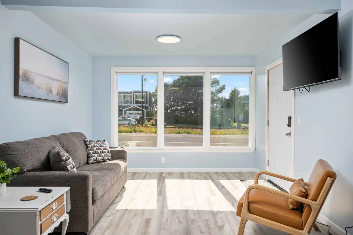 A stylish living room featuring a plush sofa, armchair, large windows letting in sunlight, and a television. The space feels modern yet cozy, with tasteful decor and an airy ambiance, ideal for unwinding after a day at the beach.