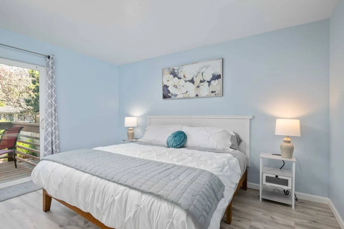 A serene bedroom with a neatly made bed adorned with crisp white linens and decorative pillows. Elegant lamps flank the bed, and a large window brings in ample natural light, creating a peaceful setting for rest or reading.