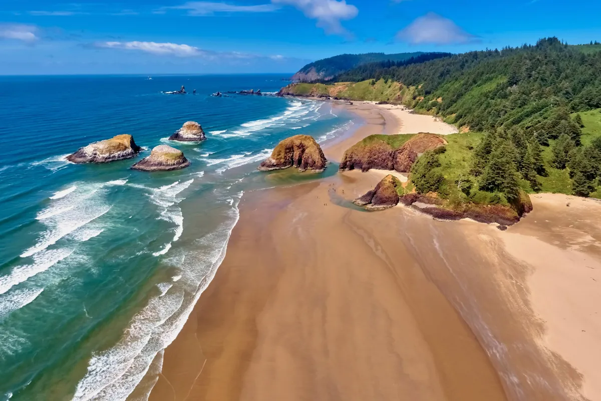 This image captures the natural beauty of the coast, showcasing dramatic rock formations, gentle waves, sandy beach, and lush greenery. The setting is tranquil yet brimming with opportunities for exploration and relaxation.