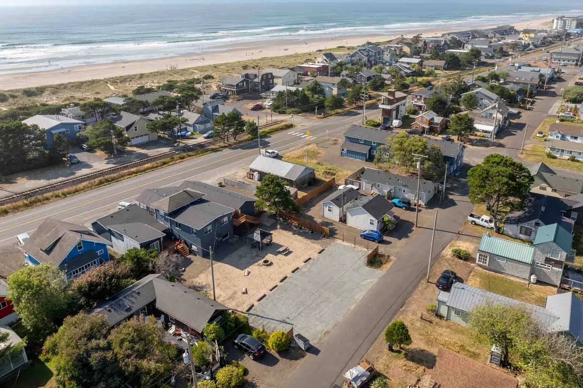 A sweeping exterior shot of Rockaway Beach at sunset, with the ocean and soft sands stretching out in the foreground. Neighborhood houses line the edges, and a roadway and greenery create a relaxed coastal vibe. The sky is tinged with warm sunset hues, suggesting the perfect setting for evening walks along the beach.