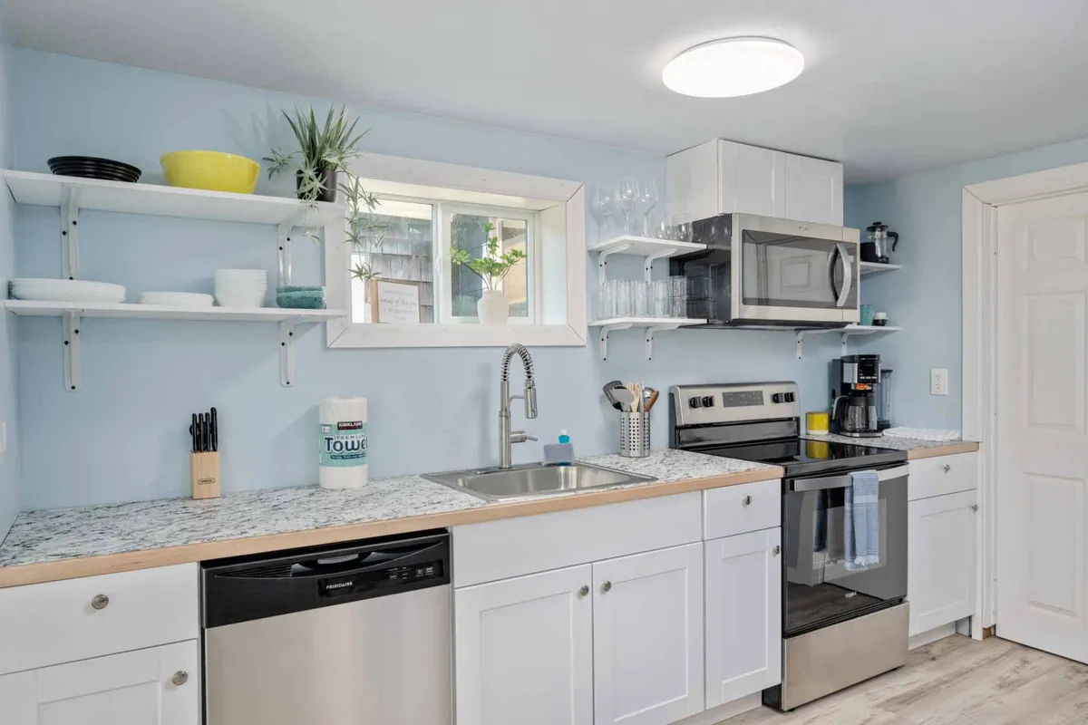 A cheerful, functional kitchenette featuring a kitchen sink, stainless steel appliances, open shelving, and a window with greenery outside. The space is bright and inviting, well-suited for cooking or light meal prep.